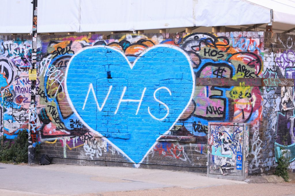 Graffiti mural with a large blue heart and 'NHS' painted on a brick wall.